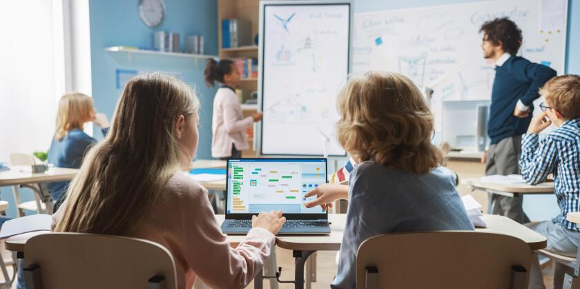 Elementary School Science Class: Over the Shoulder Little Boy and Girl Use Laptop with Screen Showing Programming Software. Physics Teacher Explains Lesson to a Diverse Class full of Smart Kids
