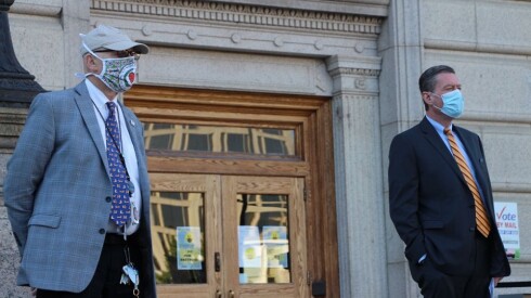 Two officials wearing facemasks stand on either side of the door to a city hall building.