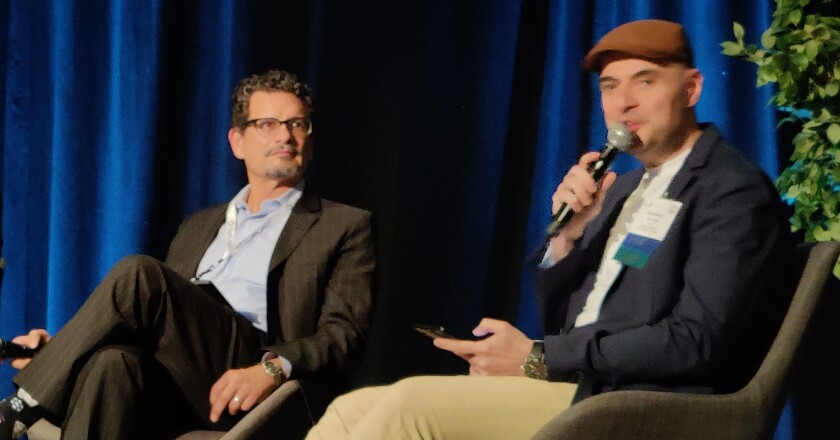 Los Angeles County Public Defender Ricardo Garcia and Chief Information Officer Mohammed Al Rawi sitting while Al Rawi is gesturing with one hand while speaking into a microphone held in the other.