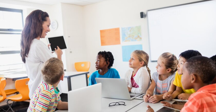 A teacher in a classroom with young students. The teacher is pointing to a tablet in her hand, and all of the students have either laptops or tablets on the tables in front of them.