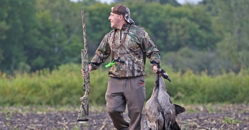A person in hunting gear carrying a rifle and two dead birds.