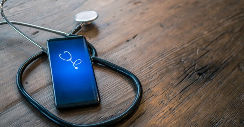 A smartphone with a white icon of a stethoscope on a blue background on its screen. The phone is lying on top of a stethoscope on a wooden surface.