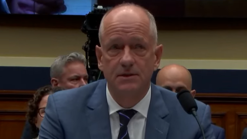 Andrew Witty, in suit and tie, seated before a microphone, reading from prepared remarks.