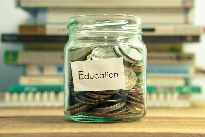 Closeup of coins in a jar that says "education" on it sitting on a wooden surface. There is a blurred stack of books in the background.