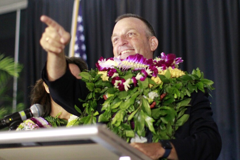 Josh Green standing at a podium holding a bouquet while smiling and pointing.