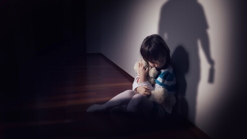 Scared child sits in corner hugging a stuffed animal.