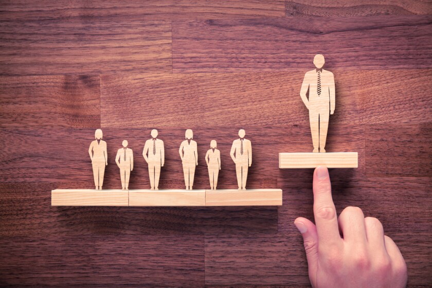 A row of wooden figures of business people with a hand pushing the largest one on the far right up higher than the rest. Dark wood background.