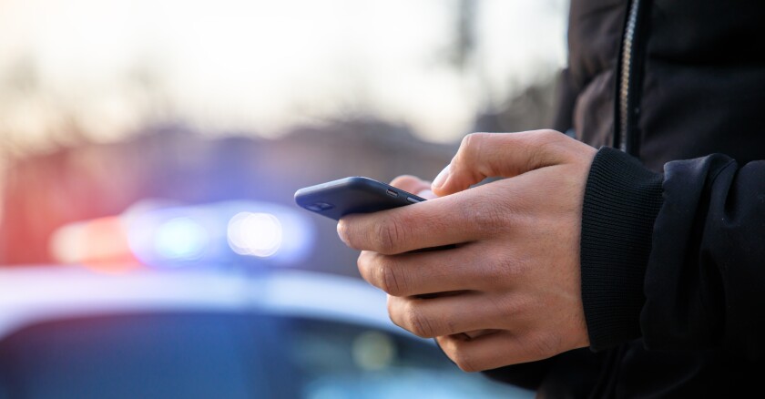Man,Holding,Phone,With,Police,Car,In,Street