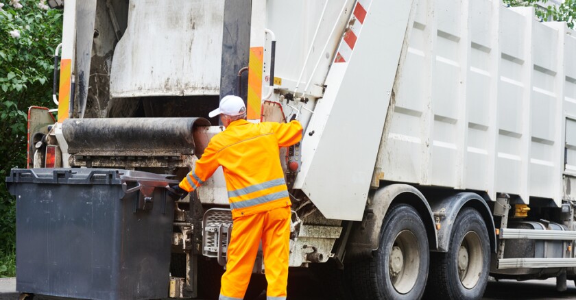 A person in yellow positioning a small dumpster to be emptied into a garbage truck.