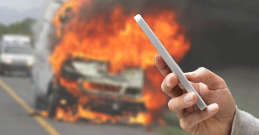 A person holding up a smartphone in the foreground. In the background, a crashed vehicle is burning on a road.