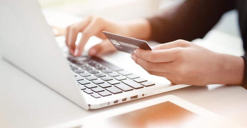 A person typing on a laptop with one hand while holding a credit card in the other.