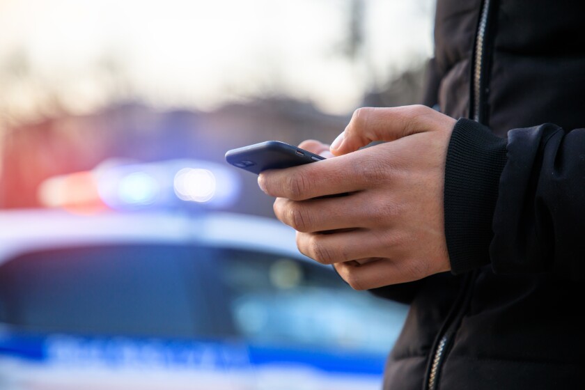 Man,Holding,Phone,With,Police,Car,In,Street
