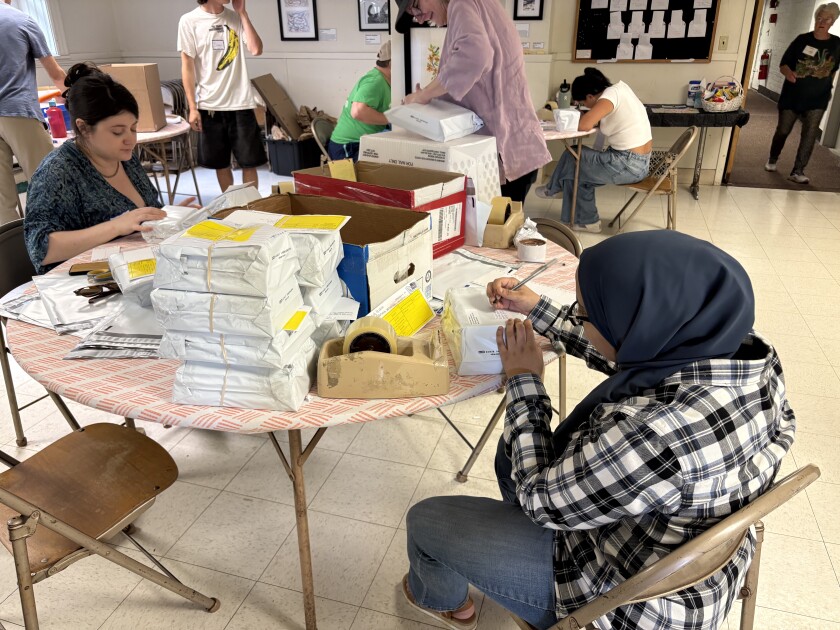 Volunteers seated around a table, where stacks of books in plastic packaging envelopes are piled high. One volunteer writes the address on a package. Another seals up an envelope.