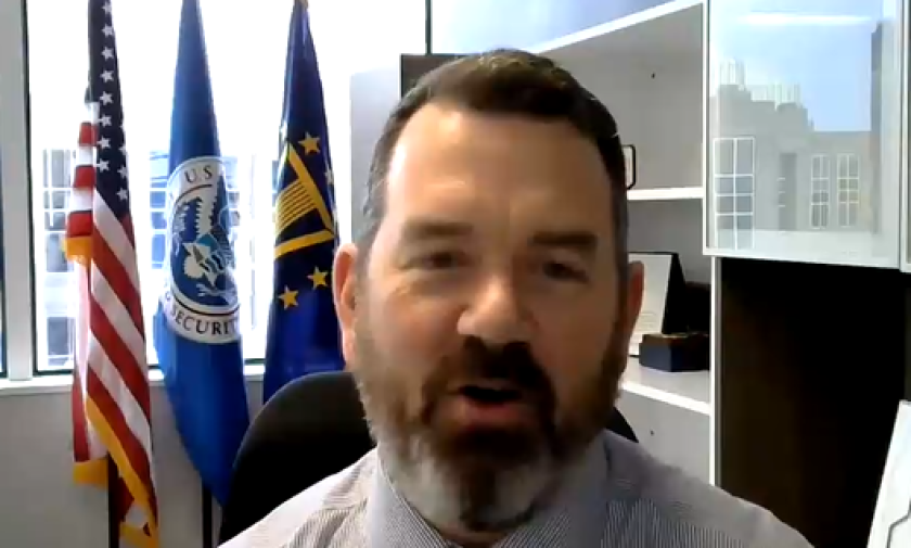 Screen shot of Trent Frazier's head and shoulders as he looks at the camera. He's in an office and flags for CISA and the USA are visible in the background.