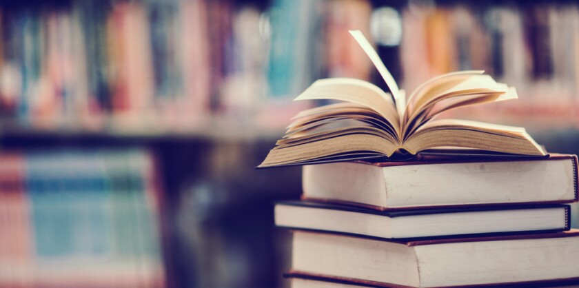 A stack of books with one open book on top of it, with a blurred shelf of books in the background.