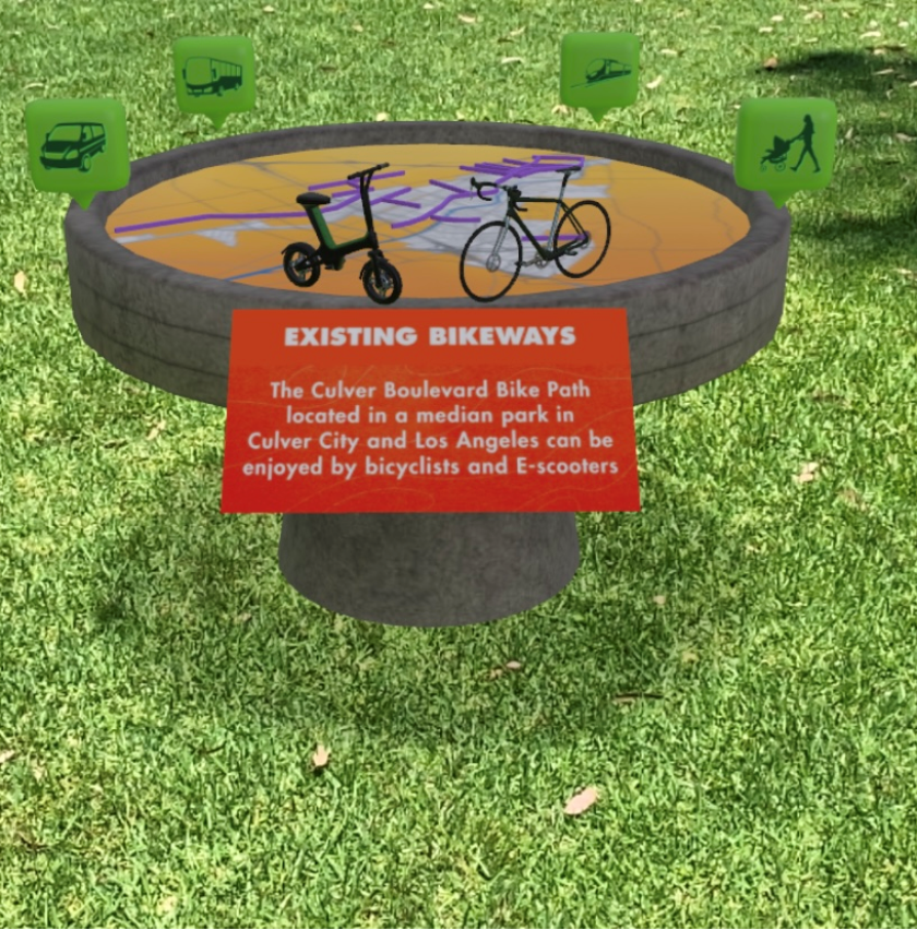 Image shows a Culver City augmented reality exhibit about mobility in the city over grass. The text reads “EXISTING BIKEWAYS” and “The Culver Boulevard Bike Path located in a median park in Culver City and Los Angeles can be enjoyed by bicyclists and E-scooters.”
