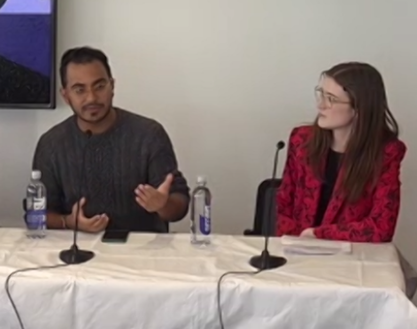 Nasser Eledroos gestures as he talks. Meg Foster turns toward him. Both are seated behind a table with microphones.