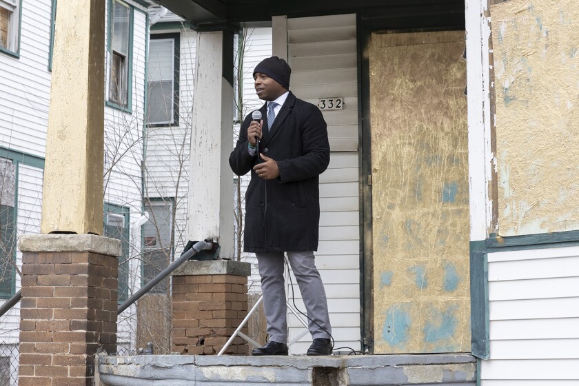 Milwaukee Mayor Cavalier Johnson standing on the porch of a blighted property speaking into a microphone.