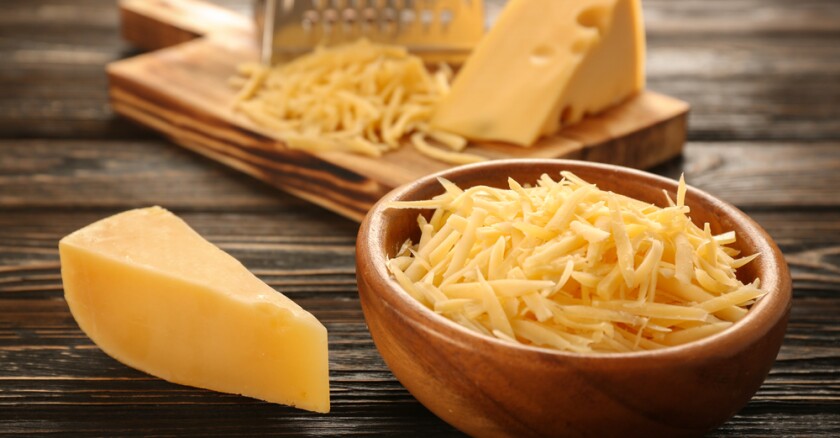 A bowl of grated cheese on a wooden table next to a large slice of cheese, with more slices and shredded cheese on a cutting board in the background.