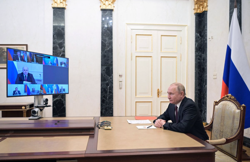 Russian President Vladimir Putin sitting at a large table in front of a screen, participating in a virtual meeting.