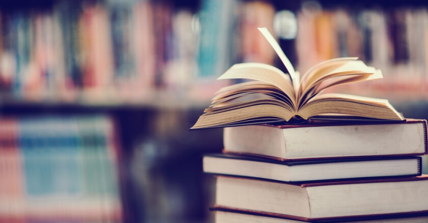A stack of books with one open book on top of it, with a blurred shelf of books in the background.