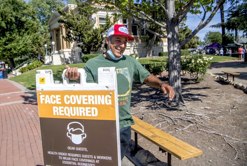 The director of a farmer’s market in Livermore, Calif., wearing a face mask pulled down to uncover his face and carrying a sign saying that masking and social distancing is required.