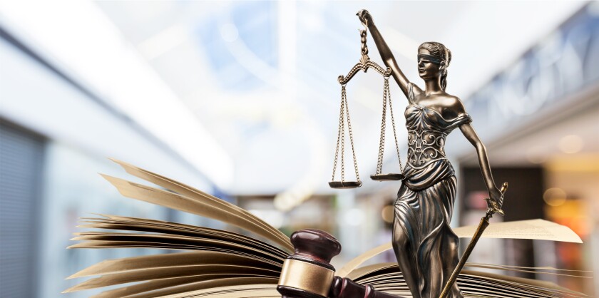 A wooden gavel resting in its pedestal with a bronze statue of Lady Justice behind it and an open book behind her. Blurry, light-colored background.
