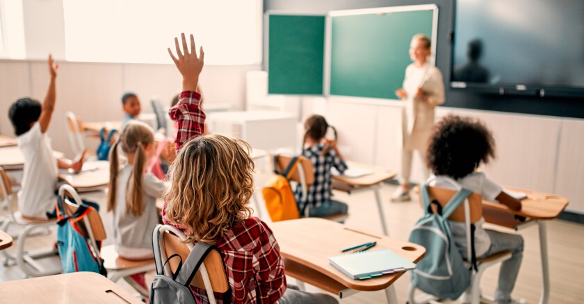 elementary school classroom with students raising their hands from their seats and a teacher at the front