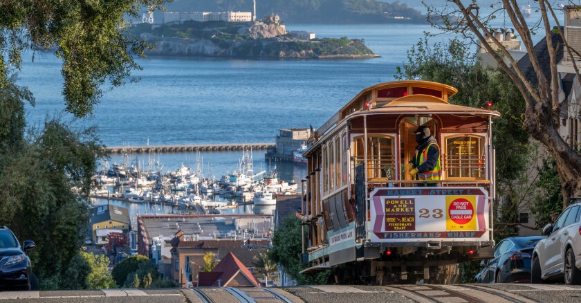 A cable car at the top of a hill about to head down in San Francisco, with Alcatraz Island and the bay visible in the background.