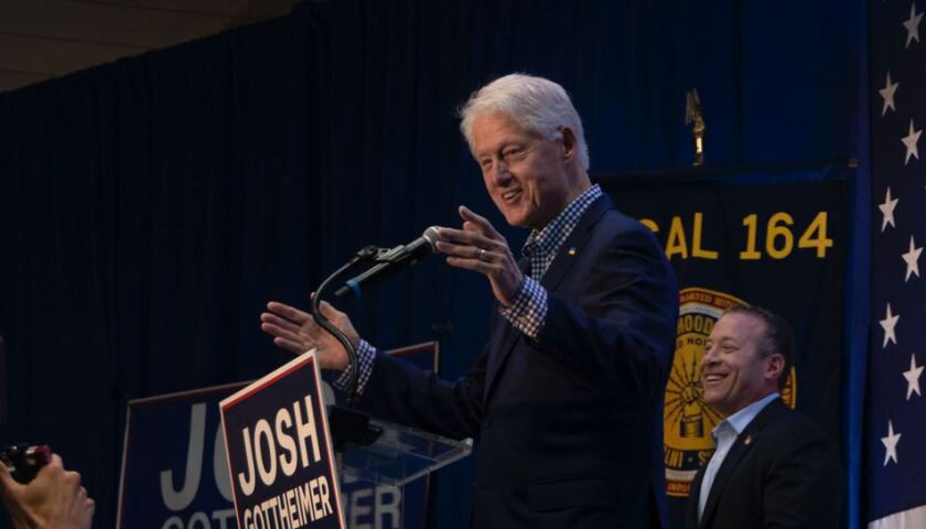 Bill Clinton smiling and gesturing with both arms while speaking into a microphone.