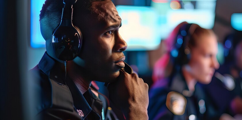 A 911 dispatcher seated in a dispatch center wearing a headset and looking at a screen that is off camera. More dispatchers are seen in the blurred background.