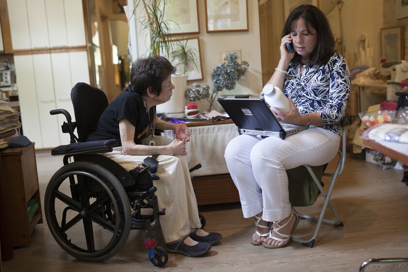 One elderly woman sitting in a wheelchair and one sitting in a folding chair talking on the phone while looking at a large pill bottle.