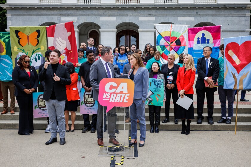 California Civil Rights Department officials and other stakeholders come together for a May 4 press conference to launch CA vs Hate. Officials stand behind podium that says CA vs Hate. People in the background hold artwork in support of effort. 