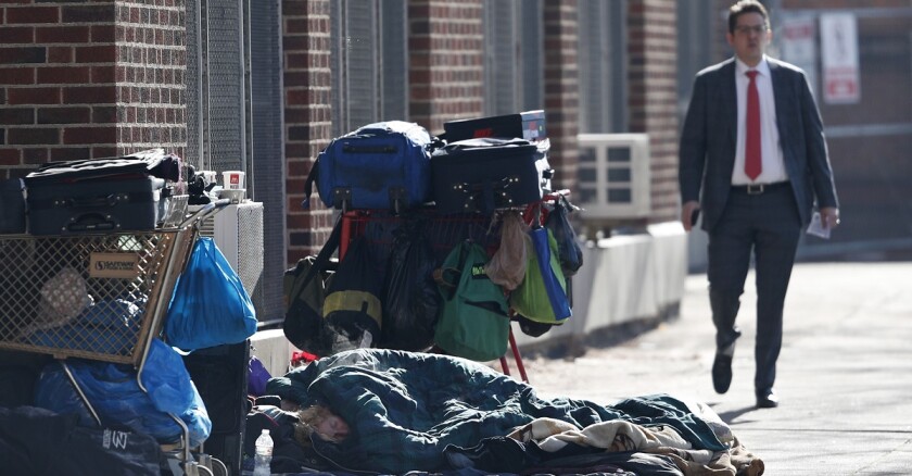 A man walking by a homeless person bundled in blankets while sleeping on the sidewalk.