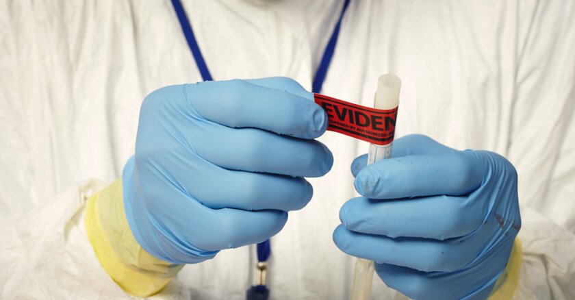 A person wearing a white full-body protective suit and blue rubber gloves putting a red "evidence" sticker on a small vial.